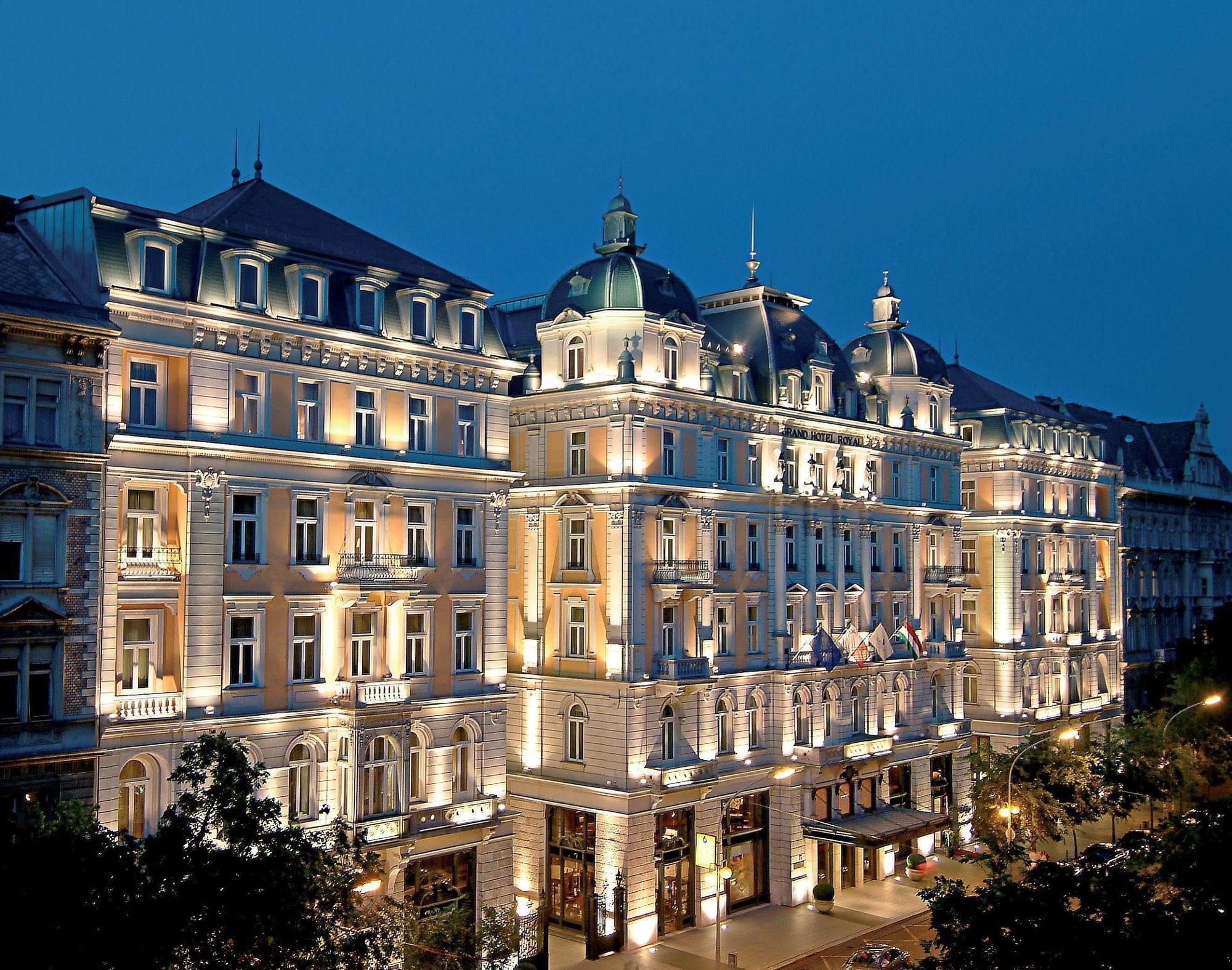 Corinthia Budapest Eksteriør billede The photo shows a large, elegantly designed building illuminated at dusk. It features architectural elements typical of classic European styles, such as ornate detailing, balconies, and a prominent roofline with domes. The facade is beautifully lit, 